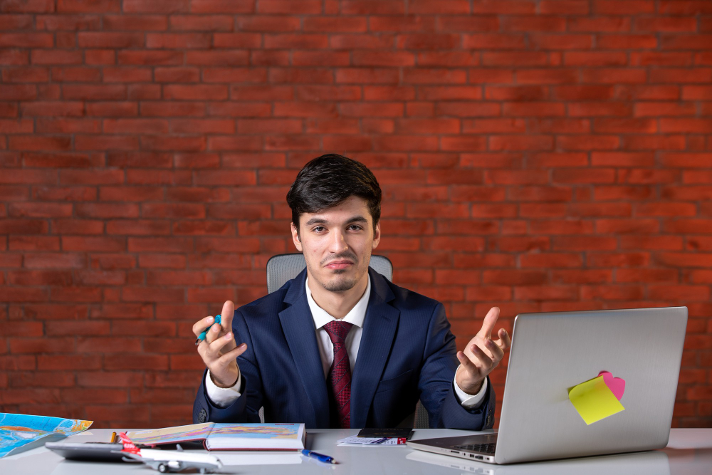 Young businessman in a suit sitting at a desk with a laptop, books, and maps, gesturing with his hands against a red brick wall background.