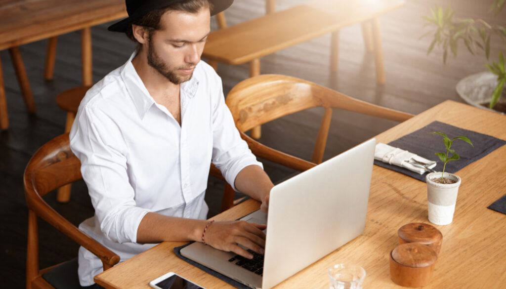 Young man wearing a hat and white shirt working on a laptop in a modern cafe with wooden furniture.