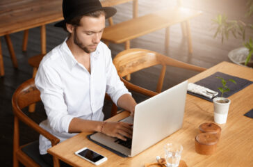 Young man wearing a hat and white shirt working on a laptop in a modern cafe with wooden furniture.