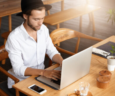 Young man wearing a hat and white shirt working on a laptop in a modern cafe with wooden furniture.